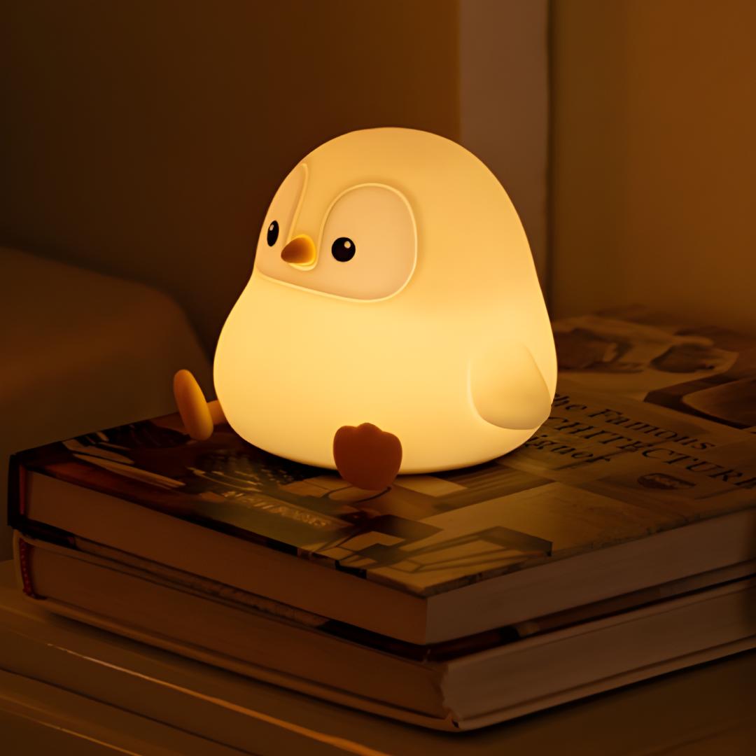 veilleuse originale en forme de pingouin assis allumée et posée sur une pile de livres à côté d'un lit