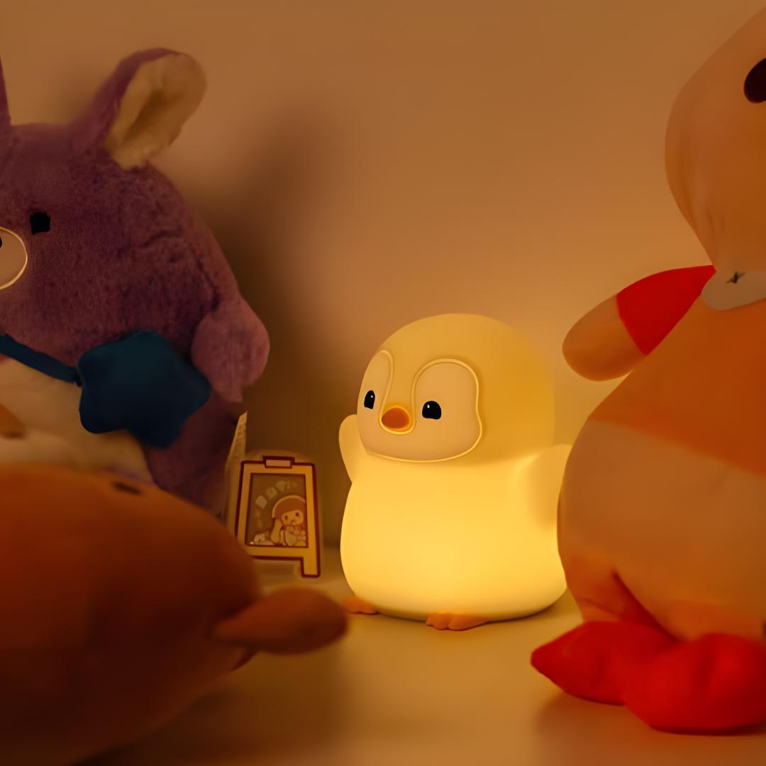 veilleuse pingouin debout avec les bras en l'air allumé et posée contre un mur avec d'autres jouets et peluches