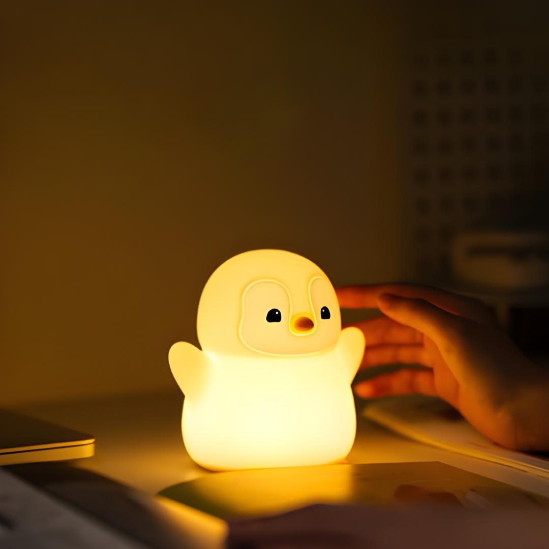 veilleuse pingouin debout avec les bras en l'air allumé et posé sur un bureau et avec une main sur le côté