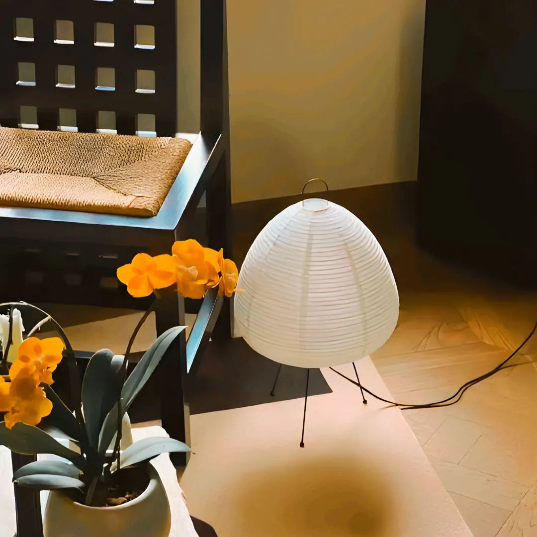 Lampe à poser en papier style japonais avec trépied noir, déco chaleureuse près d’une chaise tressée et orchidées