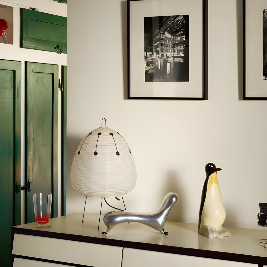 lampe en papier akari noguchi 1AD à motif sur commode blanche et avec bibelot en tout genre, photographes dans des cadres aux murs