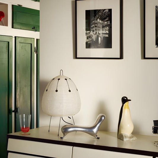 lampe en papier akari noguchi 1AD à motif sur commode blanche et avec bibelot en tout genre, photographes dans des cadres aux murs