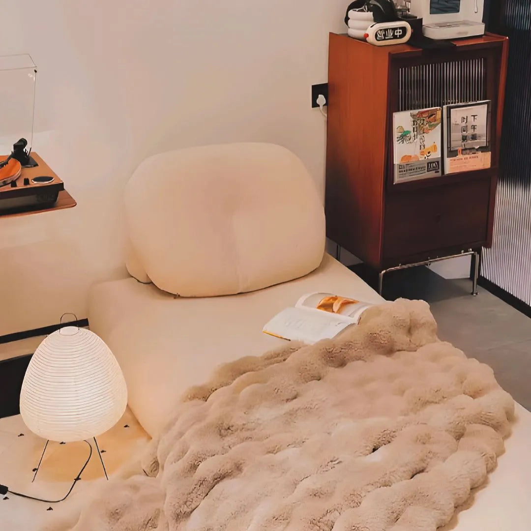 Lampe de sol en papier blanc près d’un canapé cosy, ambiance détente avec plaid et livre ouvert