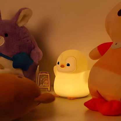 veilleuse pingouin debout avec les bras en l'air allumé et posée contre un mur avec d'autres jouets et peluches